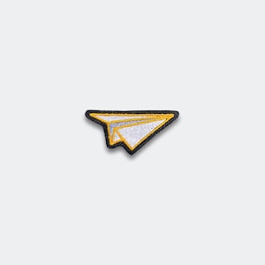 May include: Embroidered patch featuring a paper airplane design. The patch has a white paper airplane with yellow accents and a black border. This patch can be ironed on or sewn onto clothing or accessories.