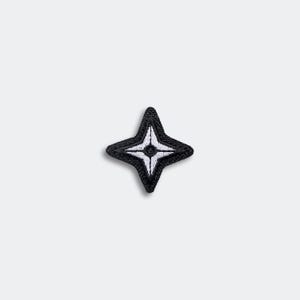 May include: A black and white embroidered patch in the shape of a four-pointed star. The patch has a black border with white detailing and a white center. This patch is suitable for clothing or accessories.