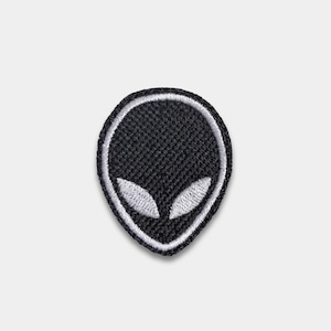 May include: A black and white embroidered patch featuring an alien head design. The alien head is oval-shaped with large, almond-shaped eyes. The patch has a white border and is suitable for clothing or accessories.