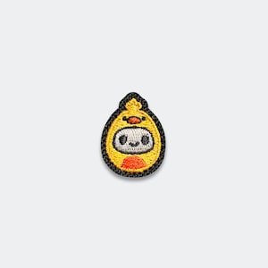 May include: An embroidered patch in a teardrop shape featuring a yellow chick costume with an orange beak and feet. The central character has a white face with black eyes and a small smile, all outlined in a thick black border on a white background.