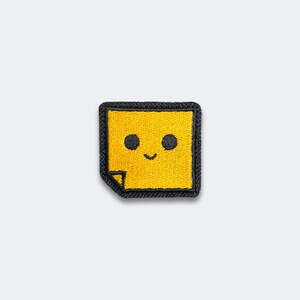 May include: Embroidered patch featuring a smiling yellow square with black eyes and a curved mouth. The patch has a black border and a small folded corner. The design is simple and cheerful, suitable for clothing or accessories.