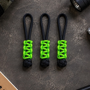 May include: Three zipper pulls made of black and neon green paracord. Each pull features a black cord loop at the top, a central section with a woven neon green pattern, and a solid black knotted ball at the bottom end of the cord.