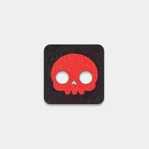 May include: A square-shaped patch featuring a black fabric background with a textured weave. Centered on the patch is a bright red skull graphic with two large circular eye sockets and a small triangular nose opening, all rendered in a flat design.