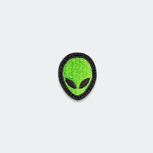 May include: An embroidered patch featuring a stylized alien head. The design consists of a bright neon green face with two large, almond-shaped black eyes, all enclosed within a thick, textured black stitched border on a neutral background.