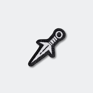 May include: A black and white embroidered patch in the shape of a kunai knife. The patch features a white blade and handle with a black outline and background. The patch is likely for clothing or accessories.