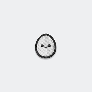 May include: An oval-shaped embroidered patch featuring a white center with a simple black stitched face consisting of two small circular eyes and a curved mouth, all enclosed within a thick, solid black stitched border edge.