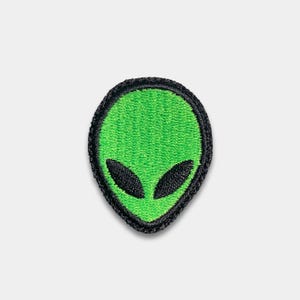 May include: A green alien patch with black eyes and a black border. The embroidered patch has a textured appearance and is in the shape of an oval. The alien's eyes are oval-shaped and black.
