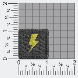 以下が含まれることがあります： 中央に黄色の稲妻のアイコンが刺繍された正方形の黒いワッペン。ワッペンはグリッド背景の上に配置されており、メートル法の定規で幅と高さが約2.5cm x 2.5cmであることが示されています。