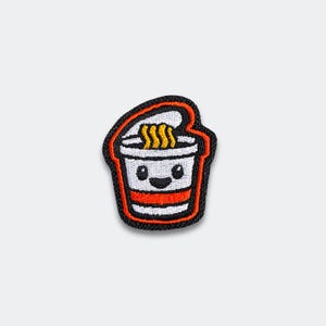 May include: Embroidered patch featuring a smiling cartoon ramen cup. The patch has a black border with an orange outline. The ramen cup is white with a red stripe and a smiling face. The noodles are yellow.