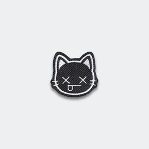 May include: Black and white embroidered patch featuring a cat's head with crossed-out eyes and a tongue sticking out. The patch has a black background with white stitching around the edges and details.
