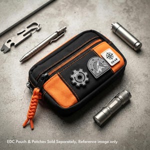 May include: A black and orange rectangular zippered pouch featuring a gear-shaped patch, a mountain compass patch, and a square patch with a compass icon and the text GEAR. The pouch has an orange paracord zipper pull, accompanied by a metal pen, a multi-tool, and two metal cylinders.