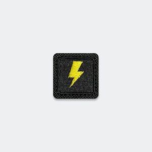 May include: A square-shaped embroidered patch featuring a black background with a bright yellow lightning bolt symbol centered in the middle. The patch has rounded corners and a thick, textured black stitched border surrounding the entire design.