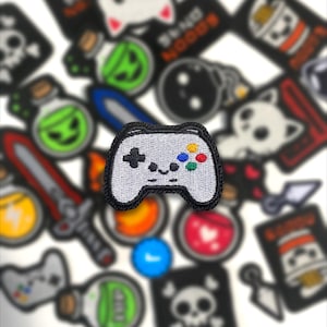 May include: A white embroidered patch of a video game controller with a black outline. The controller has a black cross, and colored buttons in blue, green, red, and yellow. Other patches with various designs are in the background.