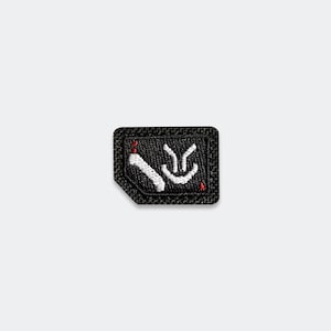 May include: A small, rectangular black embroidered patch with a clipped corner. The design features white stitched lines forming a stylized graphic symbol, accented with two small red dots at the top and bottom of the white embroidery.