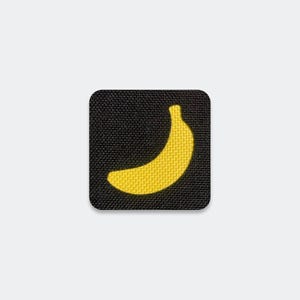 May include: A square-shaped fabric patch with a black background and a yellow embroidered banana icon centered in the middle. The patch features rounded corners and a visible woven texture throughout the entire surface of the black fabric.