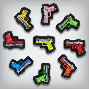 May include: A collection of embroidered patches in the shape of handguns. The patches are in various colors, including yellow, pink, green, red, blue, and orange, with black borders and silver details. The patches are arranged on a light gray background.
