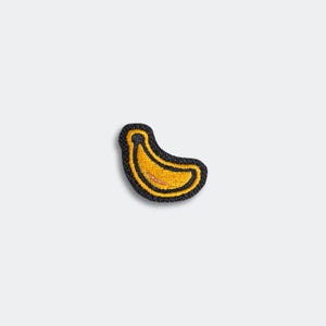 May include: Embroidered patch featuring a yellow banana with a black outline and a black border. The banana has a small pink detail. The patch is likely for clothing or accessories.