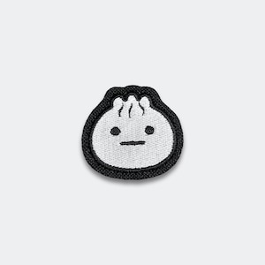 May include: Embroidered patch featuring a cartoon dumpling with a black outline. The dumpling has a white body, black eyes, and a small, horizontal black mouth. The patch is designed to be ironed or sewn onto fabric.