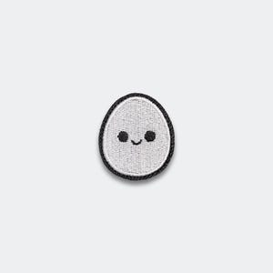May include: Embroidered patch featuring a smiling egg design. The egg is white with a black outline and a black smiling face. This patch is suitable for jackets, bags, or other fabric items.