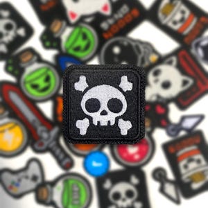 May include: Black square patch featuring a white skull and crossbones design. The patch is surrounded by other patches with various cartoon graphics, including a potion bottle, a sword, and a cat.