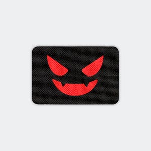 May include: A black rectangular patch featuring a red, stylized face with sharp, angled eyes and a toothy grin. The patch has rounded corners and a textured surface, creating a bold, graphic design.