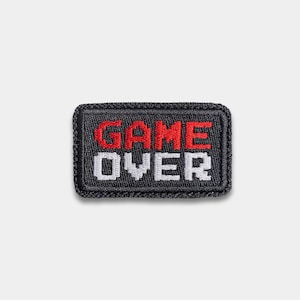 May include: A rectangular, black patch with the words "GAME OVER" embroidered in red and white pixelated letters. The patch has a black border and is likely designed to be sewn onto clothing or accessories.