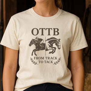 May include: A cream-colored t-shirt featuring a graphic of two horses with a jockey and the text "OTTB FROM TRACK TO TACK". The design is in dark brown, with a laurel wreath at the bottom.