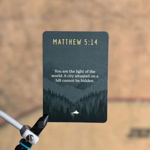 May include: A rectangular card with the text "MATTHEW 5:14" in gold lettering. The card features a dark green landscape illustration with the quote "You are the light of the world. A city situated on a hill cannot be hidden." A fly tying tool is in the foreground.
