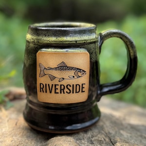 May include: A black ceramic mug with a fish illustration and the word "RIVERSIDE" on a tan square. The mug has a dark green rim and handle. The mug is sitting on a rock.