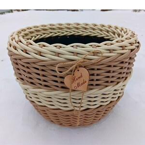 Puede incluir: Una cesta redonda tejida en tonos beige y marrón, con una etiqueta en forma de corazón que dice "Hand Made". La cesta está hecha de tiras entrelazadas, con bandas alternas de colores claros y oscuros. Un artículo decorativo.