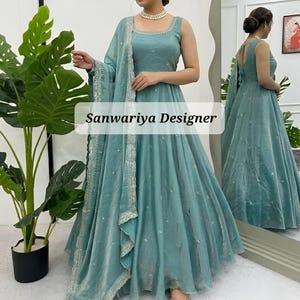May include: A light teal sleeveless gown with a matching shawl. The gown has a square neckline and a flowing skirt. The shawl and gown are decorated with silver embroidery. The text "Sanwariya Designer" is displayed on the image.