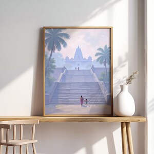 May include: Framed art print depicting a temple with a grand staircase, palm trees, and figures ascending. The artwork is in a gold frame and rests on a wooden surface. A white vase with dried plants sits nearby, adding a touch of decor.
