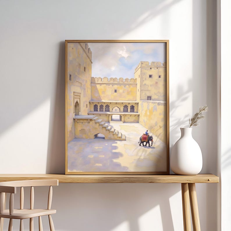 May include: A framed print of an Indian palace courtyard. The artwork depicts a yellow-toned building with a person riding an elephant. The print is displayed on a wooden table with a white vase and a sprig of dried grass.