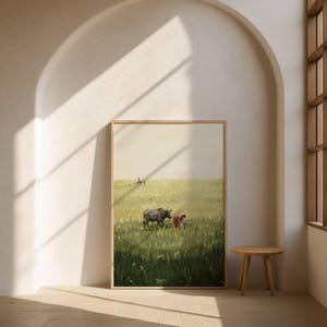 May include: A framed art print depicting a rural scene. A dark gray water buffalo stands in a field of green and yellow grass. A person in a red shirt is working in the field. The artwork is displayed in a light-colored room with a small wooden table.