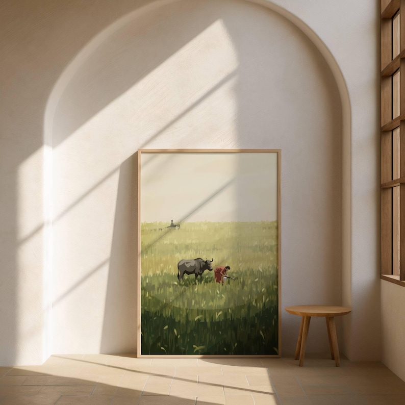 May include: A framed art print depicting a rural scene. A dark gray water buffalo stands in a field of green and yellow grass. A person in a red shirt is working in the field. The artwork is displayed in a light-colored room with a small wooden table.