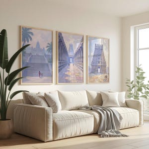 May include: Three framed watercolor prints depicting ancient temples in shades of blue, purple, and beige. The prints hang above a cream-colored sofa with several pillows and a gray throw blanket. A large potted plant is on the left.