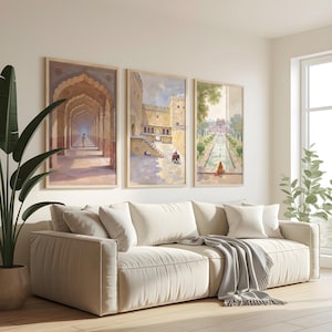 May include: Three framed landscape paintings depicting Indian architecture hang above a cream-colored sofa. The paintings feature detailed scenes of archways, buildings, and gardens. A gray throw blanket and several white pillows adorn the sofa. A large potted plant is on the left.