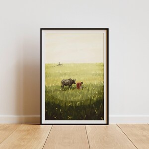 May include: Framed art print depicting a rural scene with a water buffalo and a person in a field. The artwork features a color palette of greens, browns, and yellows, with a black frame. The print is displayed on a wooden floor.