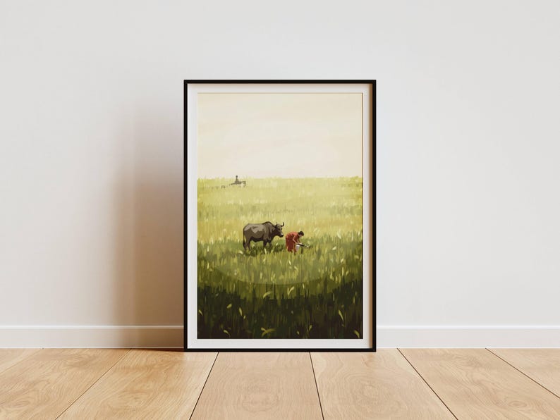 May include: Framed art print depicting a rural scene with a water buffalo and a person in a field. The artwork features a color palette of greens, browns, and yellows, with a black frame. The print is displayed on a wooden floor.