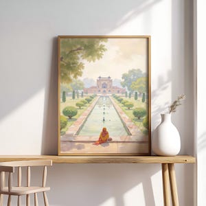 May include: A framed watercolor print depicts a serene scene of a long water feature leading to a grand building. A person in orange clothing sits near the water. The artwork is displayed on a wooden table with a white vase and chair.