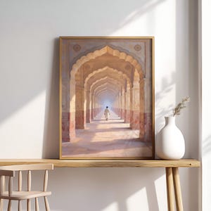 May include: Framed art print depicting a long corridor with arched ceilings and columns, leading to a distant figure. The artwork features warm, earthy tones and a sense of depth, with a gold frame. A white vase with a sprig of dried grass sits on a wooden table.