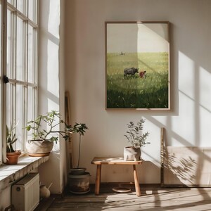 May include: A framed art print depicting a pastoral scene with a person and a dark animal in a field of tall green grass. The artwork is displayed on a white wall, with natural light streaming in from a nearby window, illuminating the room and the plants on the windowsill.