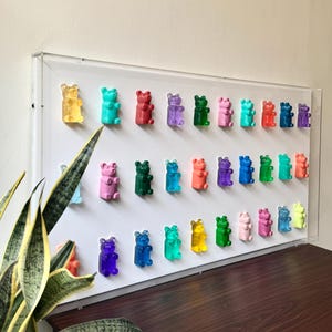 May include: A display of colourful gummy bear magnets in shades of yellow, blue, red, and green, mounted on a white surface within a clear acrylic frame. The bears are evenly spaced, creating a playful, decorative arrangement.