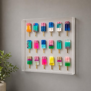 Könnte beinhalten: Ein gerahmtes Kunstwerk mit einer Sammlung farbenfroher Eis am Stiel-Skulpturen. Jedes Eis am Stiel hat eine einzigartige Farbkombination, darunter Orange, Blau, Rosa und Grün, mit dekorativen Streuseln. Die Eis am Stiel sind auf einem weißen Hintergrund montiert.