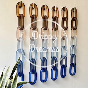 May include: Six vertical wall-mounted decorative chains made of translucent acrylic links in a gradient color scheme. Each chain transitions from amber at the top to dark blue at the bottom, suspended from gold-toned hooks. Text reads Drippy Dreamss.