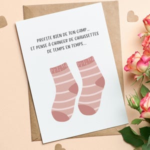 Op de afbeelding: Een wenskaart met de tekst "PROFITE BIEN DE TON CAMP... ET PENSE A CHANGER DE CHAUSSETTES DE TEMPS EN TEMPS..." en een paar roze gestreepte sokken. De kaart ligt op een bruine envelop, met hartvormige confetti en rozen.
