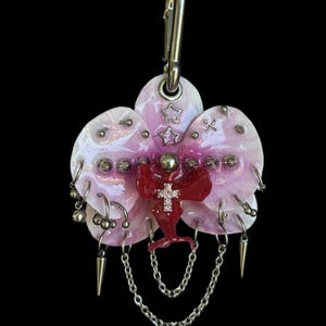 Puede incluir: Un llavero decorativo con una base en forma de orquídea rosa pálido adornada con piercings de metal plateado, dijes de estrellas y una figura roja central que sostiene una pequeña cruz, con cadenas colgantes y puntas metálicas.