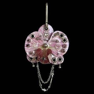 May include: A pink orchid-shaped pendant featuring a silver metal hook, multiple silver grommets, and dangling silver chains. The surface is decorated with small silver charms including a heart, an arrow, and a cross, all set against a glossy finish.