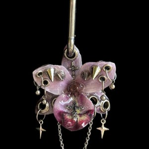 May include: A light purple orchid pendant suspended from a silver-toned metal hoop. The flower is adorned with silver-toned metal eyelets, conical spikes, small dangling ball charms, two four-pointed star charms, and two small cross charms.