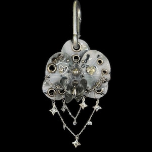 May include: A decorative charm featuring three overlapping grey and white marbled circular discs with metal grommets. A silver-toned carabiner is attached to the top. The piece is adorned with small silver chains, star-shaped charms, and heart accents.
